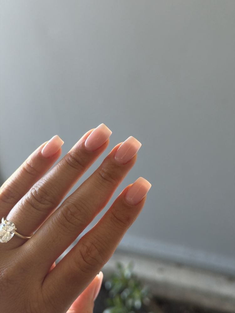 Sheer nude manicure on a hand wearing a ring, against a gray wall